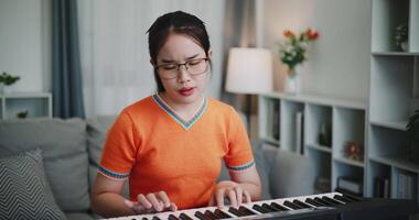 Handheld shot, Happy young woman in an orange shirt is practicing her keyboard skills intently in a living room. Enjoy favorite musical instruments video