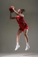 Female Basketball Player in Red Uniform Jumping for Shot during Training on Grey Studio Background photo
