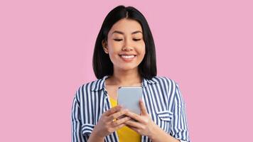 Amazing Application. Front portrait of smiling young asian woman using smartphone standing isolated over pink studio background. Cheerful lady texting sms message, chatting with friends, banner photo
