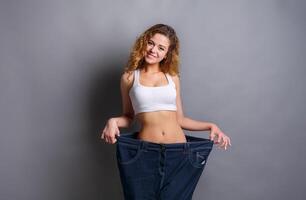 Young redhead woman wearing jeans much bigger size after loosing weight from diet, gray studio background, copy space. Healthy eating, bodybuilding concept photo