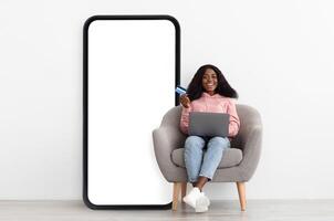 Mobile shopping application. Happy black lady sitting on chair near big cellphone with empty screen using credit card and laptop, white studio wall background, mockup, collage photo