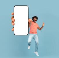 Emotional curly millennial indian guy in casual jumping up with brand new smartphone with white blank screen over blue studio background, gesturing, mockup, full length shot, copy space photo