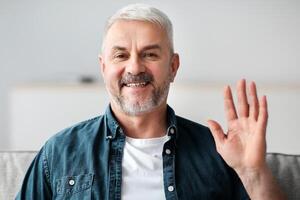 Handsome senior man having conference, smiling and waving at camera, device screen with telecommunication software frame. Elderly man having online party during COVID-19 pandemic photo