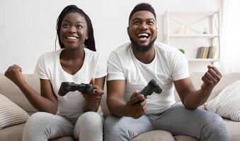 Fun hobbies for couples. Joyful black guy and girl celebrating victory in game, holding joysticks, raising fists and laughing, panorama photo