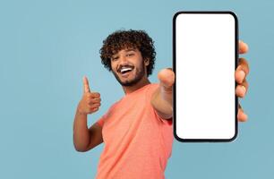 Modern smartphone with white blank screen in positive curly, bearded indian guy hand, positive eastern man showing nice mobile application and thumb up, blue background, mockup, copy space photo