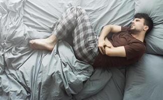 Young man suffering from stomach ache, lying in bed at home, top view. Health problems concept photo