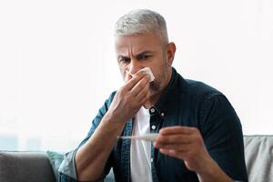 Sick senior man sneezing nose and checking body temperature with thermometer, sitting on sofa at home. Matured grey-haired man feeling sick, got coronavirus, cold or flue, copy space photo