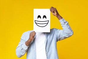Happiness and success concept. Black male hiding face behind sheet of paper with drawn happy emoticon over yellow background in studio, copy space photo