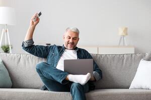 Relaxed mature man sitting on couch with laptop, turning on AC, feeling hot. Happy senior man enjoying time at home, surfing on Internet, using notebook, holding remote for air conditioner, copy space photo