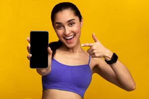 Sports App. Portrait of excited fit young lady holding and showing smartphone, pointing and indicating black empty screen for mockup template, wearing fitness tracker, orange studio background photo