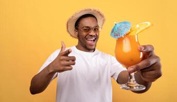 Carefree black guy in casual wear holding tropical cocktail and pointing at camera on yellow studio background. Cool young man drinking yummy alcohol-free beverage on summer vacation photo