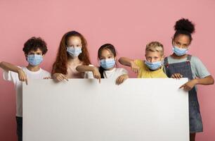 Multiethnic group of kids in medical masks pointing at blank banner over pink background, mockup for design photo