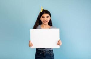 Amazing promo. Happy Indian teen girl holding blank white banner with copy space on blue studio background, mockup. Beautiful adolescent showing poster with place for your ad photo