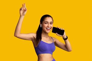 Music Lover. Portrait Of Cheerful Happy Sporty Woman Wearing Wireless Earbuds, Holding Smartphone As Mic And Singing, Listening To Favourite Playlist Isolated On Orange Studio Background photo
