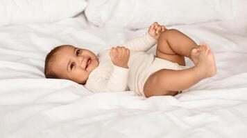 In The Morning. Portrait of adorable smiling black infant lying on the back in bed on white bedsheet at home. Positive healthy laughing afro child wearing bodysuit, looking at camera. Happy Childhood photo