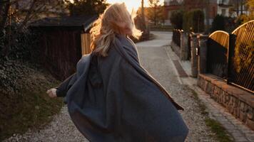 Cheerful blonde female wrapping herself in soft gray blanket, spinning freely and looking back,during golden sunset light in residential driveway with windswept golden hair and radiant smile video