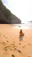 FPV drone shot of woman meditating alone on remote tropical beach with turquoise sea, waves, and cliffs, Bali, Indonesia. Peaceful scene of relaxation, mindfulness, and healing retreat. video