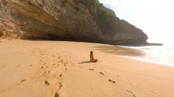 FPV drone shot of woman meditating alone on remote tropical beach with turquoise sea, waves, and cliffs, Bali, Indonesia. Peaceful scene of relaxation, mindfulness, and healing retreat. video