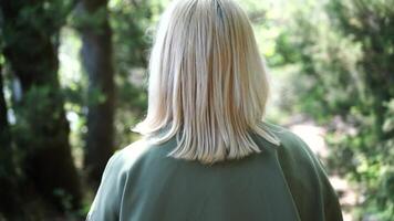 Blonde walking in the park. Happy young woman wearing. Happy young woman in a green jacket walks forward in the park, rear view, slow motion video