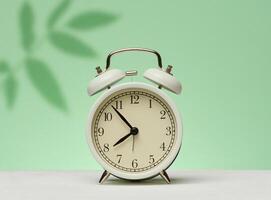 White vintage alarm clock stands on a green surface against a background with shadows from leaves. Waking up and starting the day. photo