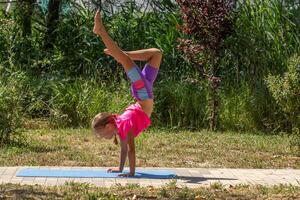 A young girl practices a dynamic handstand on a yoga mat outdoors, supporting her weight on both hands while one leg is extended upward and the other bent. photo