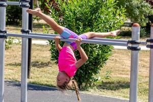 A child hangs upside down from a horizontal bar, legs extended in a full split, demonstrating impressive flexibility and body control while suspended in the air. photo