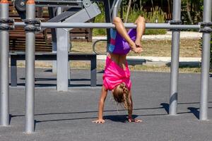 A child balances upside down on a horizontal bar, supporting the body with bent arms while the legs are lifted and bent overhead. The pose shows strength, coordination, and gymnastic control. photo