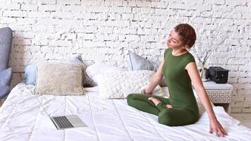 A slender woman in a green bodysuit performs yoga exercises online, sitting on a white bed video