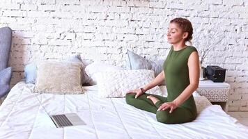 A woman performs yoga exercises online, sitting on a white bed, flexing her shoulders video