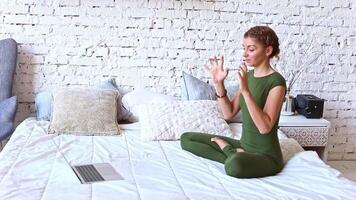 A woman performs yoga exercises online, sitting on a white bed, a laptop is in front of her video