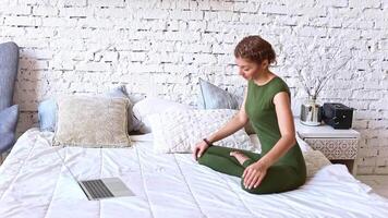A woman in a green jumpsuit performs yoga exercises online, sitting on a white bed video