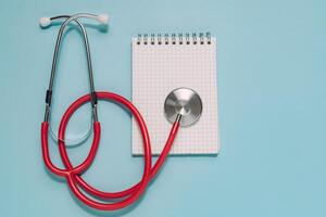 Vibrant red stethoscope next to an open spiral notepad with grid paper, all on light blue. Ideal for medical consultation, patient records, diagnosis, and healthcare planning. photo