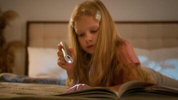 Focused blond little Caucasian girl child kid reading book pages interesting fairytale schoolgirl daughter study do homework distant learning use flashlight lying bed evening bedroom indoors apartment video
