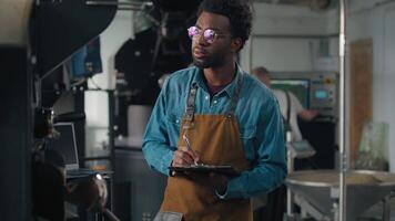 Confident smiling African American man worker in coffee roasting fabric work with roasted cocoa machine checking inspecting writing notes expertise quality control engineer professional guy working video