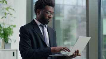 African American businessman male business man in office looking out window typing laptop smiling happy working at workplace using corporate software chatting internet browsing analyzing online data video