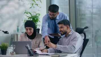 Business Arabian team three Indian colleagues in office discuss problem documents looking at computer happy celebrate work winning achieve looking at screen analyze financial growth success high five video
