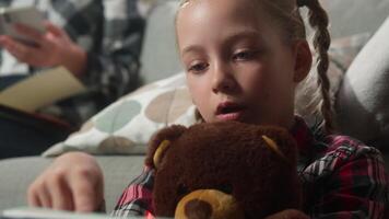 Concentrated Caucasian child girl kid daughter reading book studying homework hug teddy bear mother working with phone writing notes notebook at home background family parenting remote job freelance video