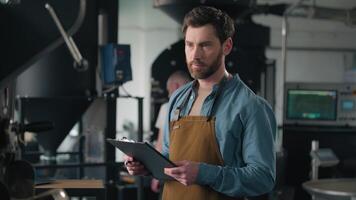 Caucasian man barista small business owner coffee roasting machine control check inspection production factory male guy writing notes roaster quality checklist thoughtful inspector manufacture worker video