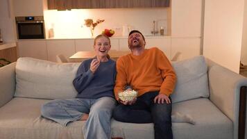 Happy joyful romantic couple laughing and eating popcorn while watching comedy show on TV at home video