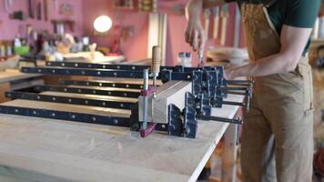 Busy carpenter employs metal clamps to fix glue boards beams together in furniture making studio video