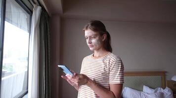 Woman with cosmetic mask on the face using smartphone and looking out of the window in hotel room , moisturizing her skin after plane journey video