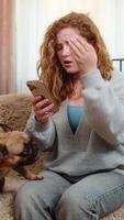 Young Caucasian redhead woman gets bad news on smartphone while sitting on home sofa, showing disappointment and tension. Girl frowns as pug dog nearby senses mood and reacts with curious posture. video