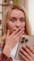 Young adult woman sits on home sofa reading surprising news on smartphone with wide eyes and curious look. Blonde girl reacts with awe wow and amazement to unexpected positive update or event online. video