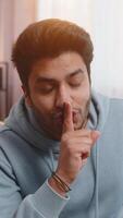 Shh be quiet please. Portrait of young Indian man presses index finger to lips makes silence gesture sign do not tells secret, stop talk gossip, confidential privacy. Guy sitting on sofa at home room. video