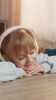 Young girl lying on table listening to an audiobook with headphones, reacting with excitement, surprise. Caucasian child smiles joyfully, wondering what happens next in story, enjoys adventure at home video