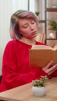 Young woman concentrated reading book sitting at home table, thoughtfully turning pages serious expression. Caucasian girl deeply involved in literature hobby demonstrating focus calm leisure activity video