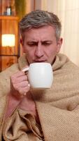 Young sad man at home sits wrapped in blanket with cup of tea, looking sick and coughing. Caucasian guy on sofa shows cold symptoms, rests quietly and tries to recover from mild illness and fatigue video