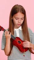 Young child girl joyfully plays a toy ukulele, pretending to perform on stage, smiling with enthusiasm musical imagination while posing on a vibrant background. School kid isolated on pink background video
