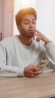 Young Indian man sitting on sofa at table, actively texting on smartphone, thoughtfully resting head on hand in living room at home. Hispanic guy looks focused, deeply engaged in conversation or task. video
