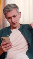 Young man at home sits on sofa, shakes head while reading smartphone and shows disappointment. Caucasian guy looks upset and frustrated after losing or getting bad news in message or social feed video